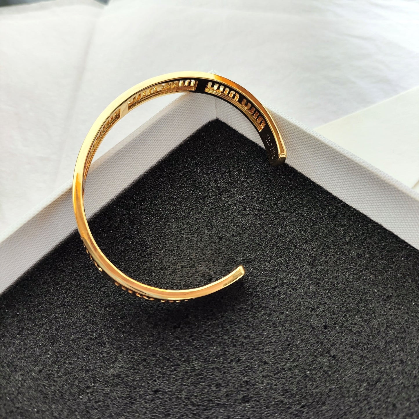 Gold open bangle cuff with engraved miumiu logo, resting on textured cushion