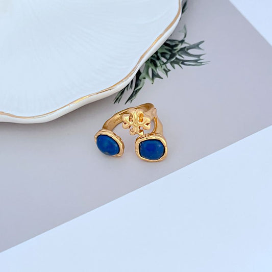 Gold-tone open metal ring with acrylic logo and blue stone accents