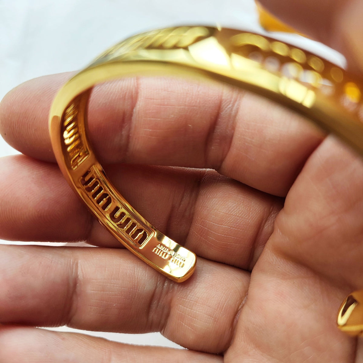 Gold metal bangle bracelet detail showing engraved “miu miu” logo on side.