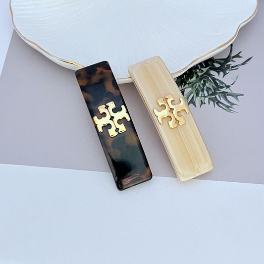 Two acrylic logo hair clips: milky white and dark tortoise with gold emblem