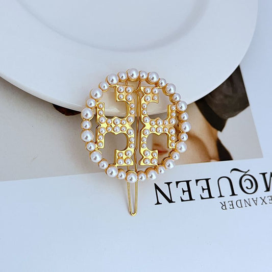 Gold metal hair clip with circular openwork logo, bordered by white pearls